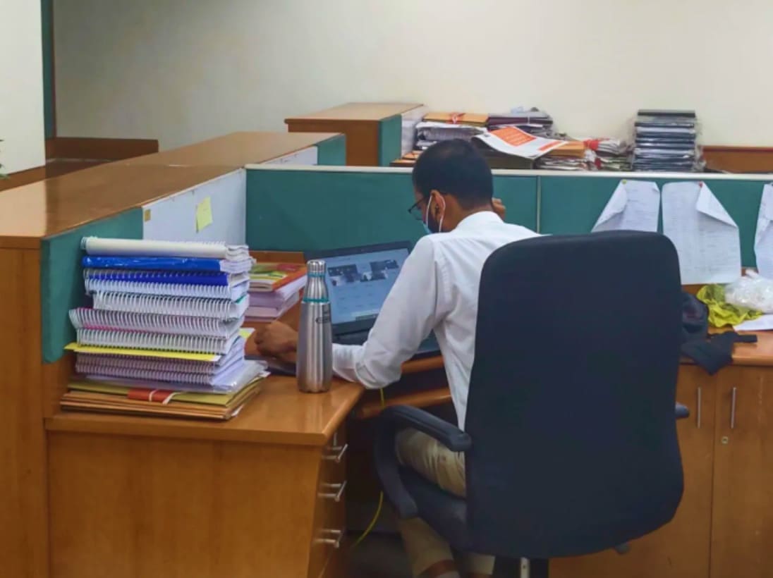 A man at a laptop in a government office cubicle, piles of files on the desk.