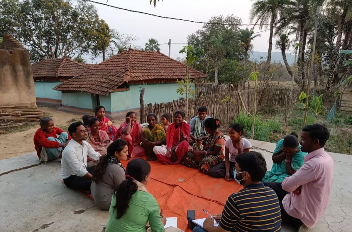 A village gathering seated outside a house, listening.